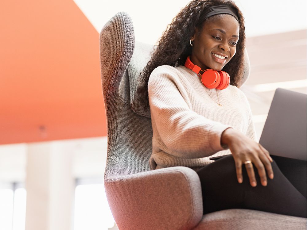 A Henkel employee sits in a comfy chair with her laptop on her knees. She smiles.