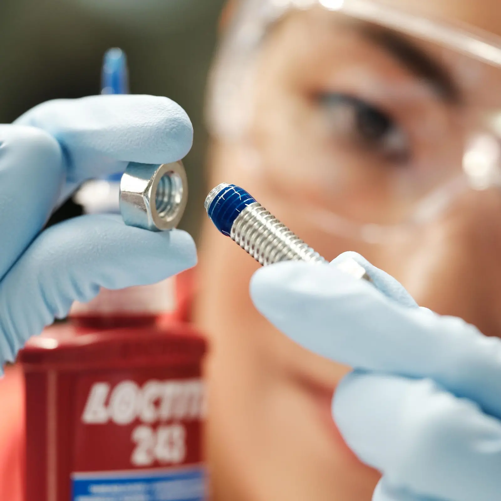 Das Gesicht einer Frau im Hintergrund. Im Vordergrund sieht man Ihre Hände in blauen Plastikhandschuhen eine Schraube mit blauer Schraubensicherung haltend.