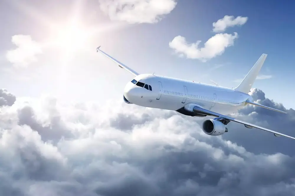 Ein weißes Passagierflugzeug fliegt über weichen Wolken in einem hellen Himmel, das Sonnenlicht scheint von links oben.