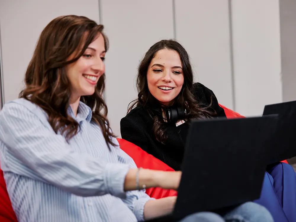 Zwei Personen sitzen in einem modernen Büro auf farbigen Sitzsäcken und arbeiten jeweils an einem Laptop. Sie scheinen sich über Benefits für Henkel-Mitarbeitende auszutauschen und kollaborativ zu arbeiten.