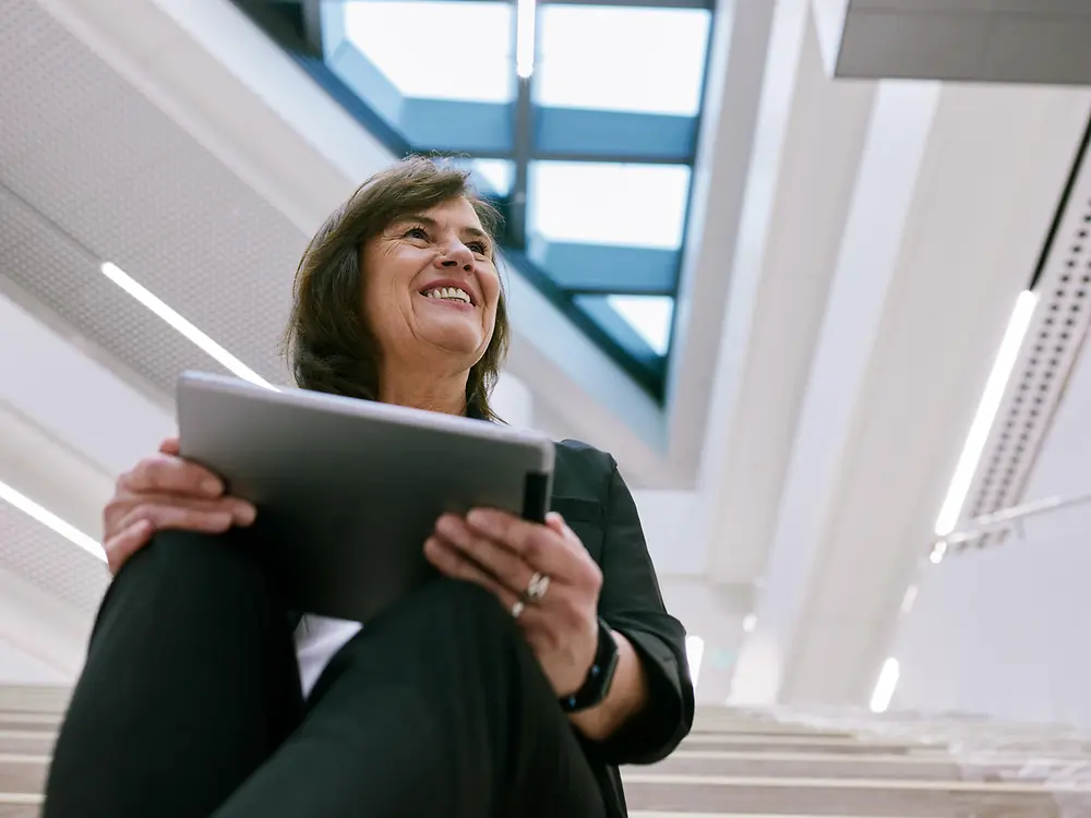 Eine Person sitzt auf einer Treppe in einem modernen Gebäude und hält ein Tablet in der Hand. Im Hintergrund ist eine helle, offene Architektur mit großen Fenstern und Oberlichtern zu sehen, die eine klare und professionelle Atmosphäre schafft.