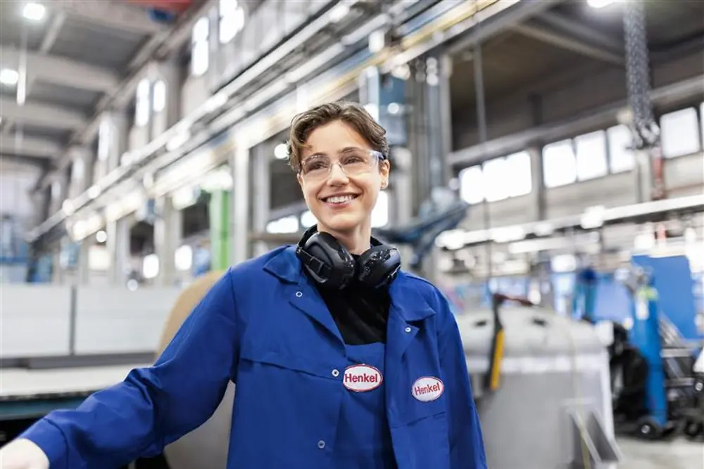 Ein lächelnder Henkel-Mitarbeiter trägt einen blauen Arbeitsmantel und Gehörschutz und steht in einer industriellen Produktionshalle – stellvertretend für Expertise und Innovation in der Fertigung.