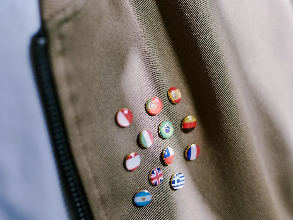 Nahaufnahme eines braunen Stoffes mit einem Reißverschluss auf der linken Seite und mehreren kleinen runden Pins mit verschiedenen Nationalflaggen – als Symbol für internationale Vielfalt.