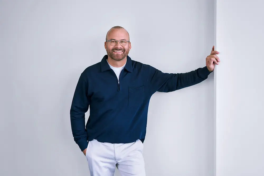 Eine berufserfahrene Person bei Henkel steht selbstbewusst vor einer weißen Wand in einem modernen Umfeld und trägt ein dunkles Langarmshirt sowie eine helle Hose.