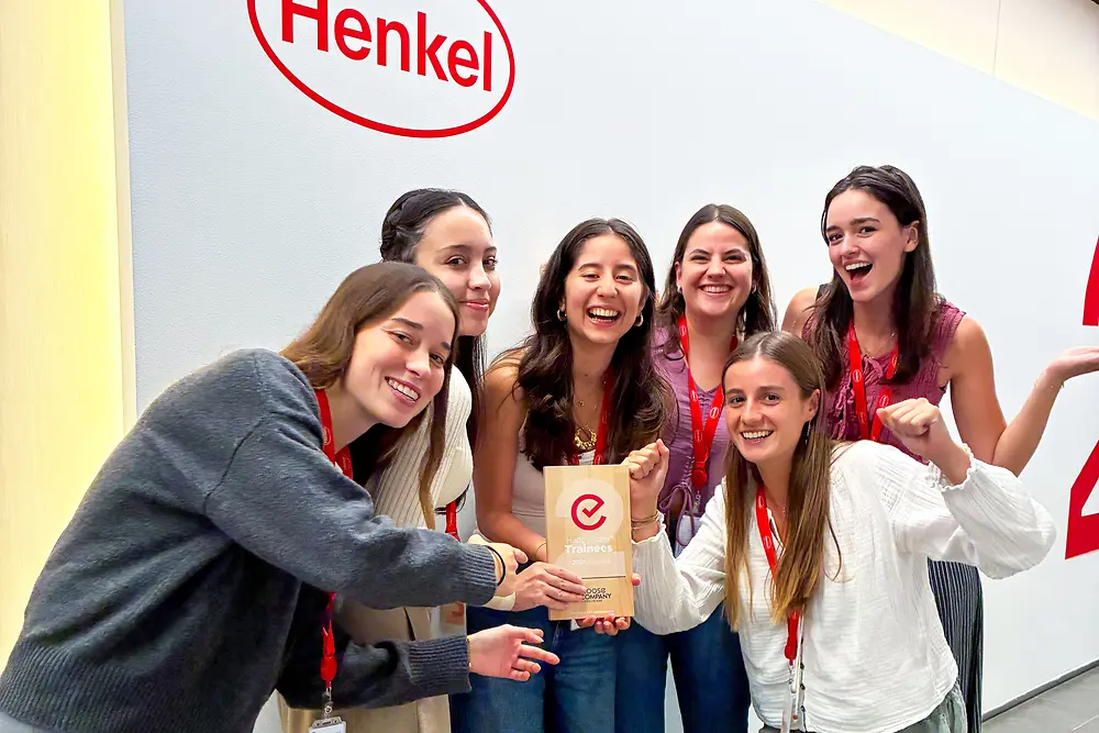 Eine Gruppe aus sechs Personen steht gemeinsam vor einer Wand mit dem Henkel-Logo. Sie tragen rote Lanyards und legere Kleidung und versammeln sich eng um eine Auszeichnungsplakette. Die Szene spielt in einem modernen Unternehmensumfeld mit heller Wand und grauem Boden. Die Gruppe posiert begeistert und hebt die Auszeichnung in der Mitte hervor.