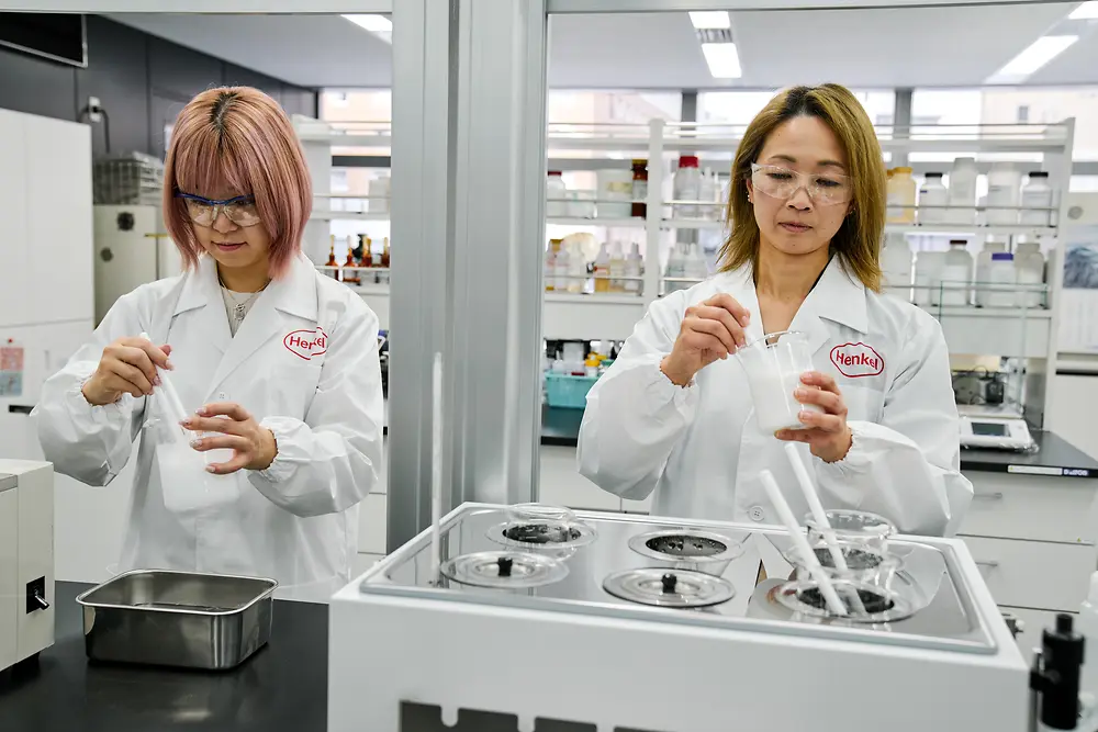Zwei Personen in weißen Henkel-Laborkitteln arbeiten in einem modernen Labor. Sie bedienen wissenschaftliche Geräte und Behälter auf einer Arbeitsfläche; im Hintergrund sind verschiedene Laborgeräte und Regale mit Materialien zu sehen.